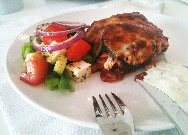 Moussaka, et kjøttstykke med saus og en hvit dressing anrettet på en hvit tallerken. Foto.