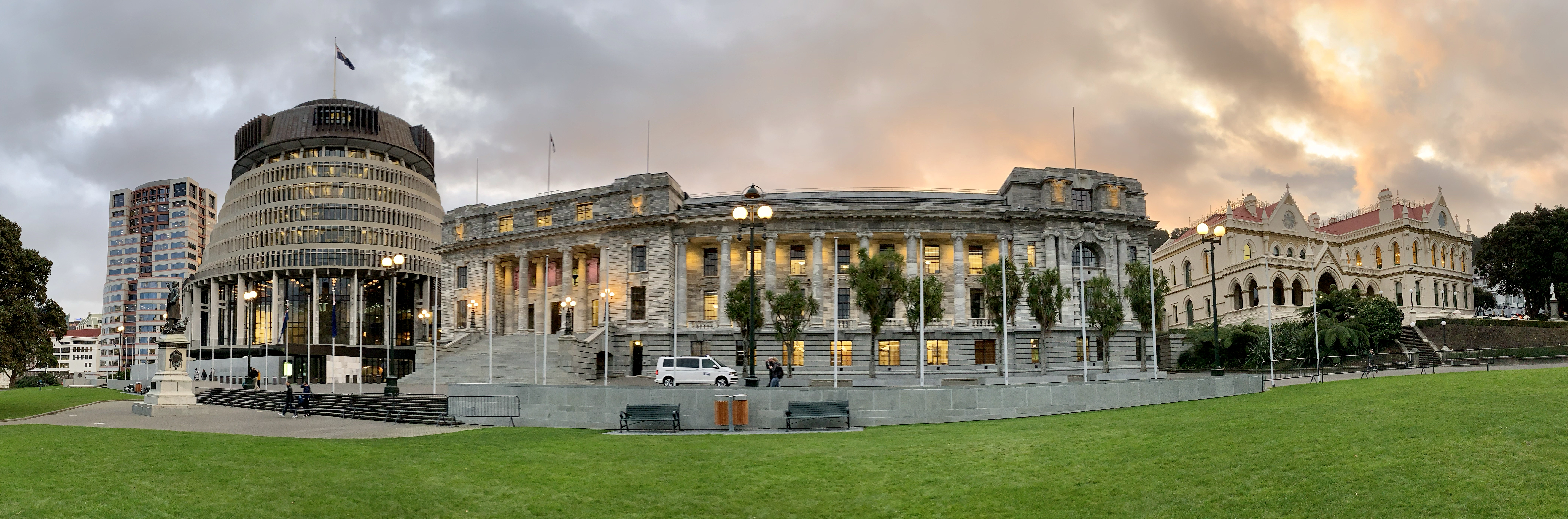 Photo: We see several buildings. To the right we see a modern building 'the Beehive' which houses the executive branch in New Zealand. To the right of it we see the parliament building, which is a Victorian style stone building. 