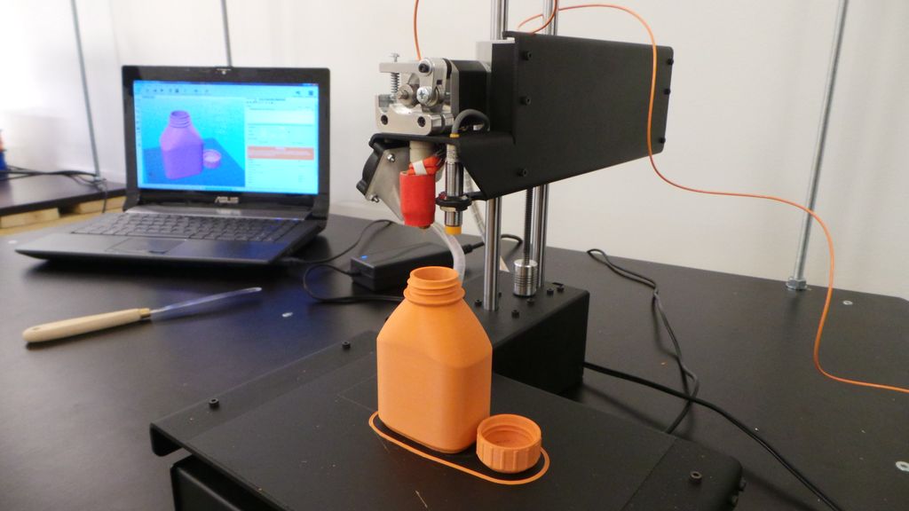 En 3D-printer lager ei flaske og en kork i plast. I bakgrunnen står en bærbar pc med programvare for 3D-printer og bilde av flaska og korken på skjermen. Foto.