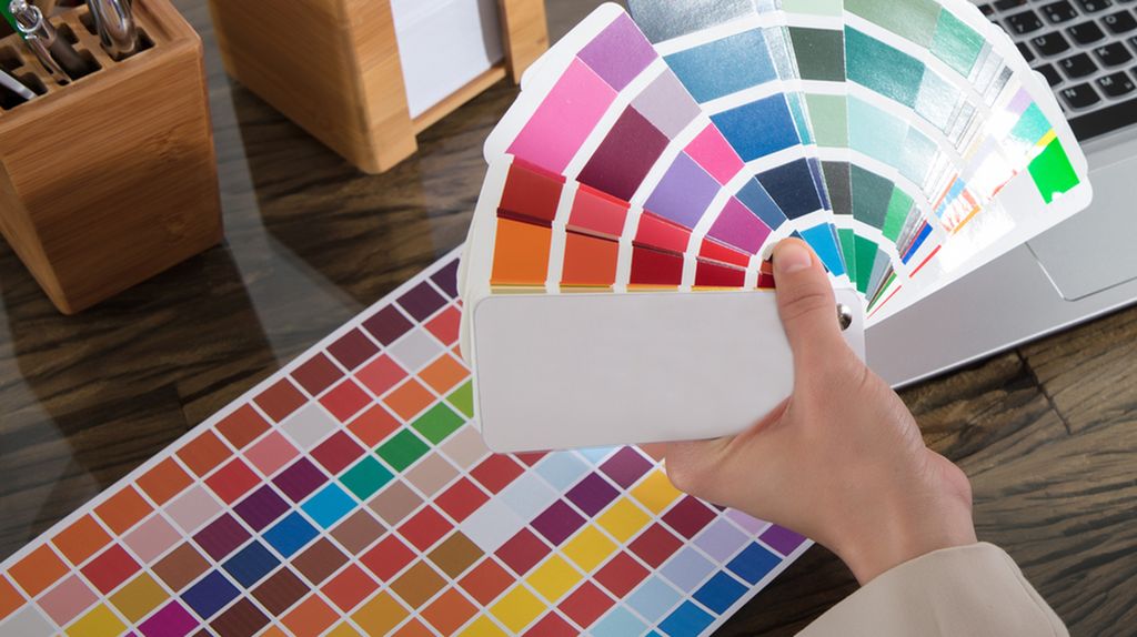 Hånd holder ei vifte med fargeprøver over et arbeidsbord med flere fargeprøver og en bærbar datamaskin. Foto.