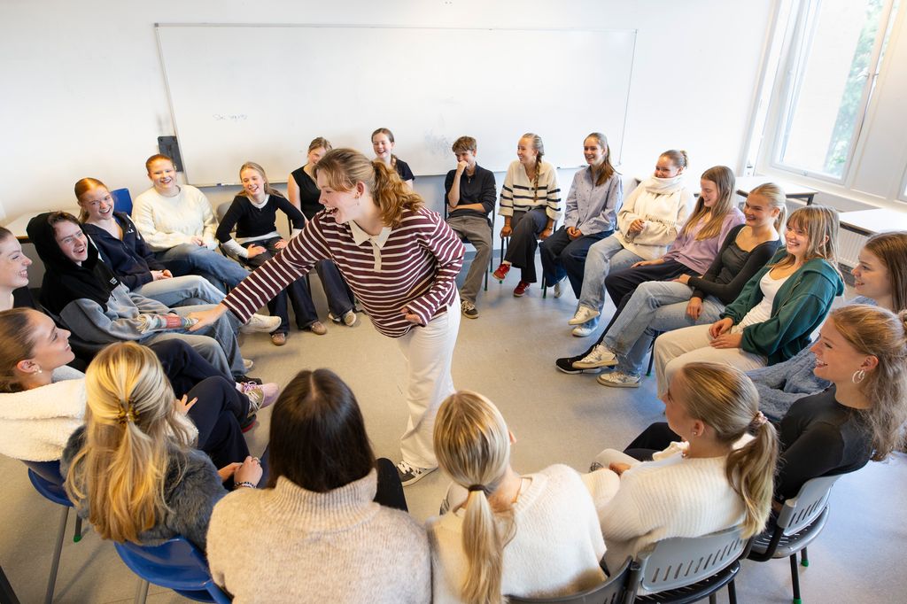 Ei stor gruppe med elever sitter i ring på stoler i et klasserom. I midten står en elev med en sammenbrettet avis og peker på en av elevene i gruppa. Alle ler og ser ut til å more seg sammen. Foto.  