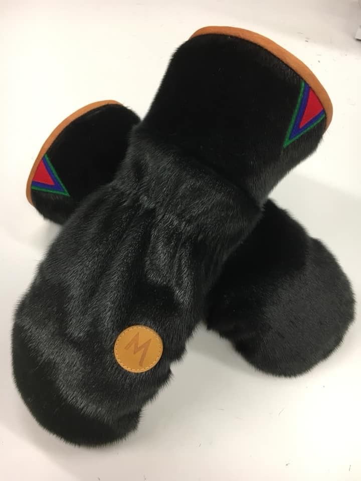 Et par votter i svart selskinn, med designmerke og samisk dekor: en okerfarget kant og  en spiss liten trekant i rødt, blått og grønt. Foto.