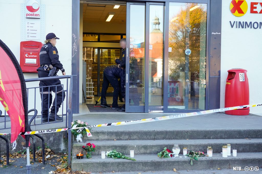 Inngangspartiet til eit butikklokale, med sperreband og politi som arbeider på staden. Blomstrar og lys er lagde på trappa til butikken. Foto.