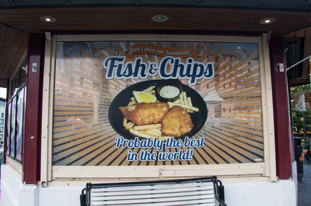 Stor plakat på en fasade i ei gågate. Teksten på plakaten er som følger: "Don't miss our homemade Fish & Chips. Probably the best in the world!" Midt på plakaten er det bilde av en porsjon med fritert fisk og pommes frites. Foto.