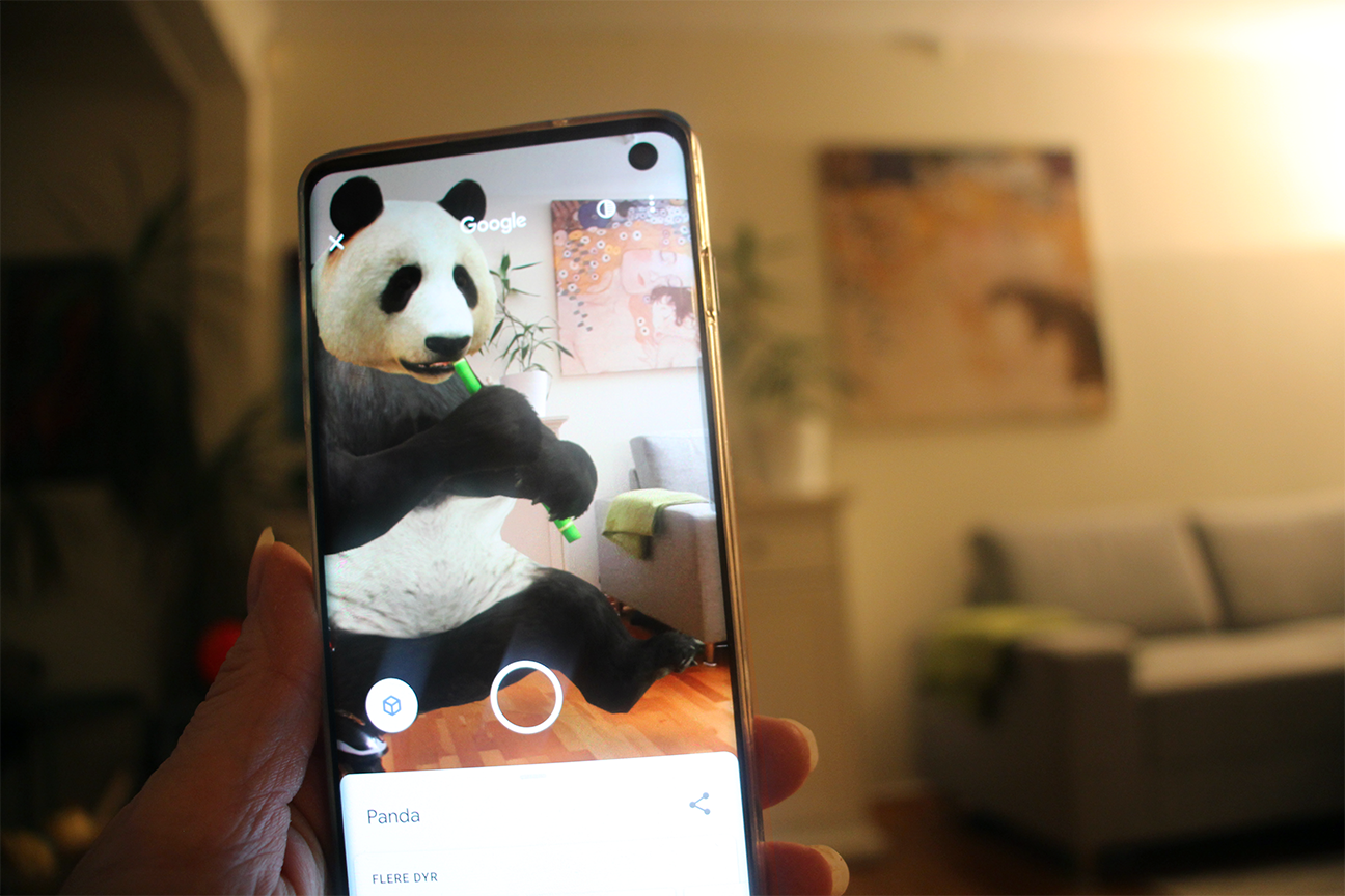 Ei hand held ein mobil framom ein sofa på ei stove. På mobilen er det ein panda som sit på golvet, ved den same sofaen. Foto.
