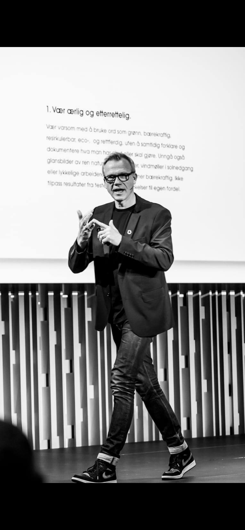 En person står foran en vegg med tekst. Han henvender seg i retning kamera og snakker mens han gestikulerer med hendene. Foto.