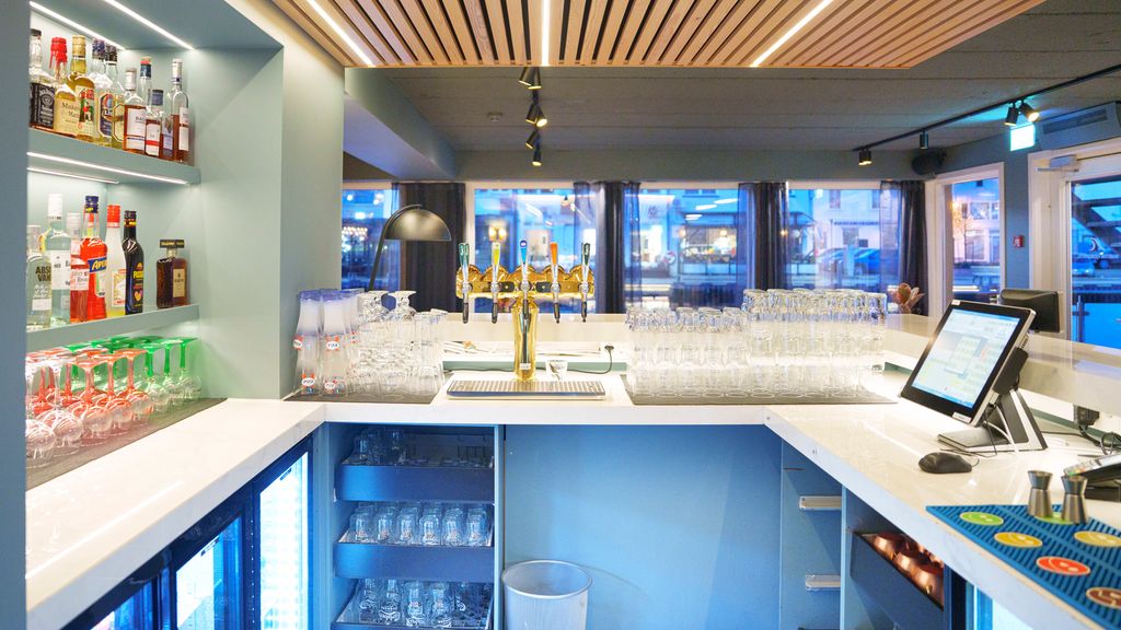 Interiørbilde fra innsiden i en bar. Bakveggen i baren har opplyste hyller med jevne rekker av flasker. På den ene siden er det et tappetårn med ulike typer rene glass i ryddige stabler på begge sider. I front er det en dataskjerm og barmatter med to målebeger. Under benkene er det lave kjøleskap med glassfront, flere rene glass på brett og ei søppelbøtte. Foto.