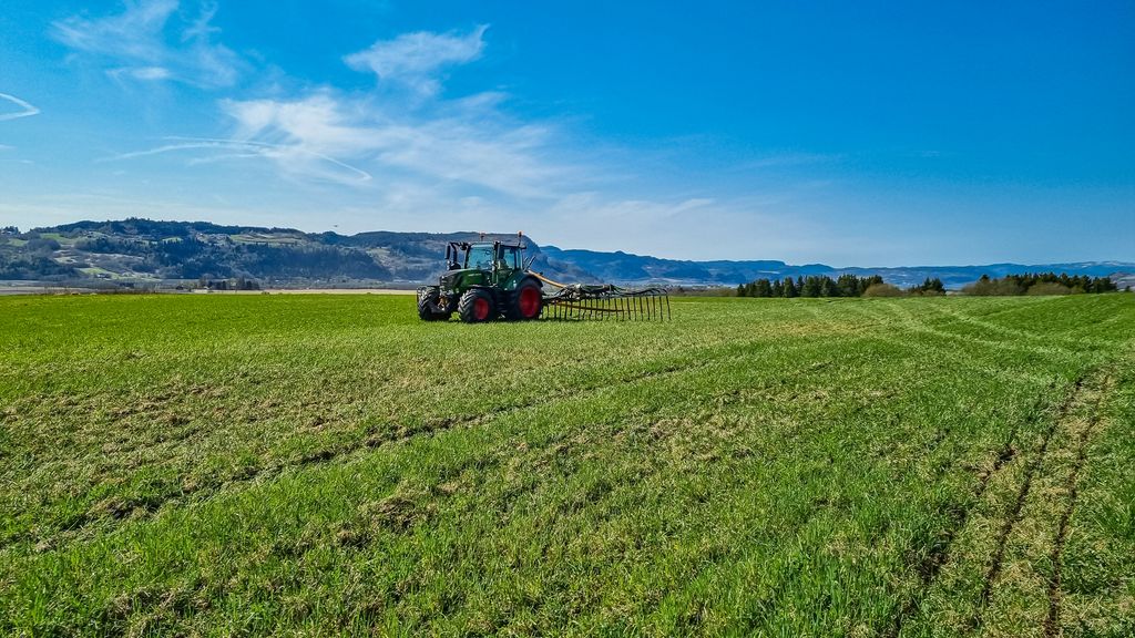Et stort grønt jorde der en traktor kjører ut husdyrgjødsel med slangespreder, med fjell og blå himmel i bakgrunnen. Foto.