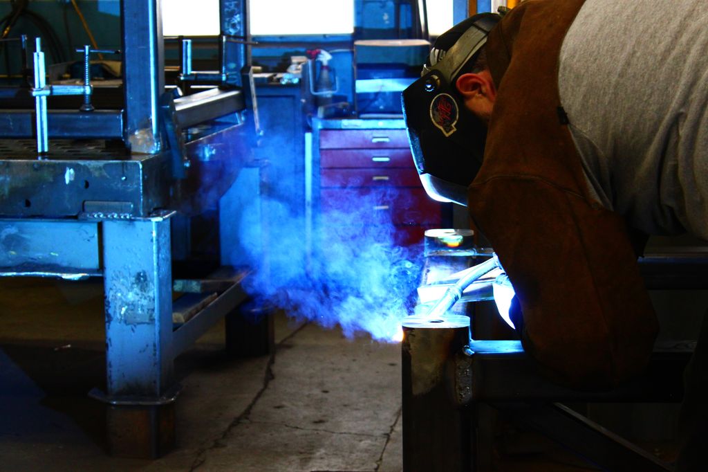 En person står bøyd over en arbeidsbenk og sveiser et objekt. Blå røyk stiger opp fra arbeidsområdet. Personen har sveisemaske. Foto.