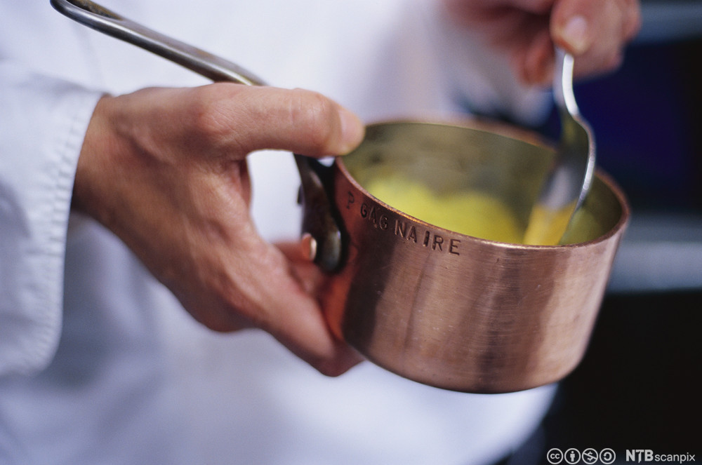 En kokk holder en liten kjele med saus i hendene og rører med ei skje. Foto.