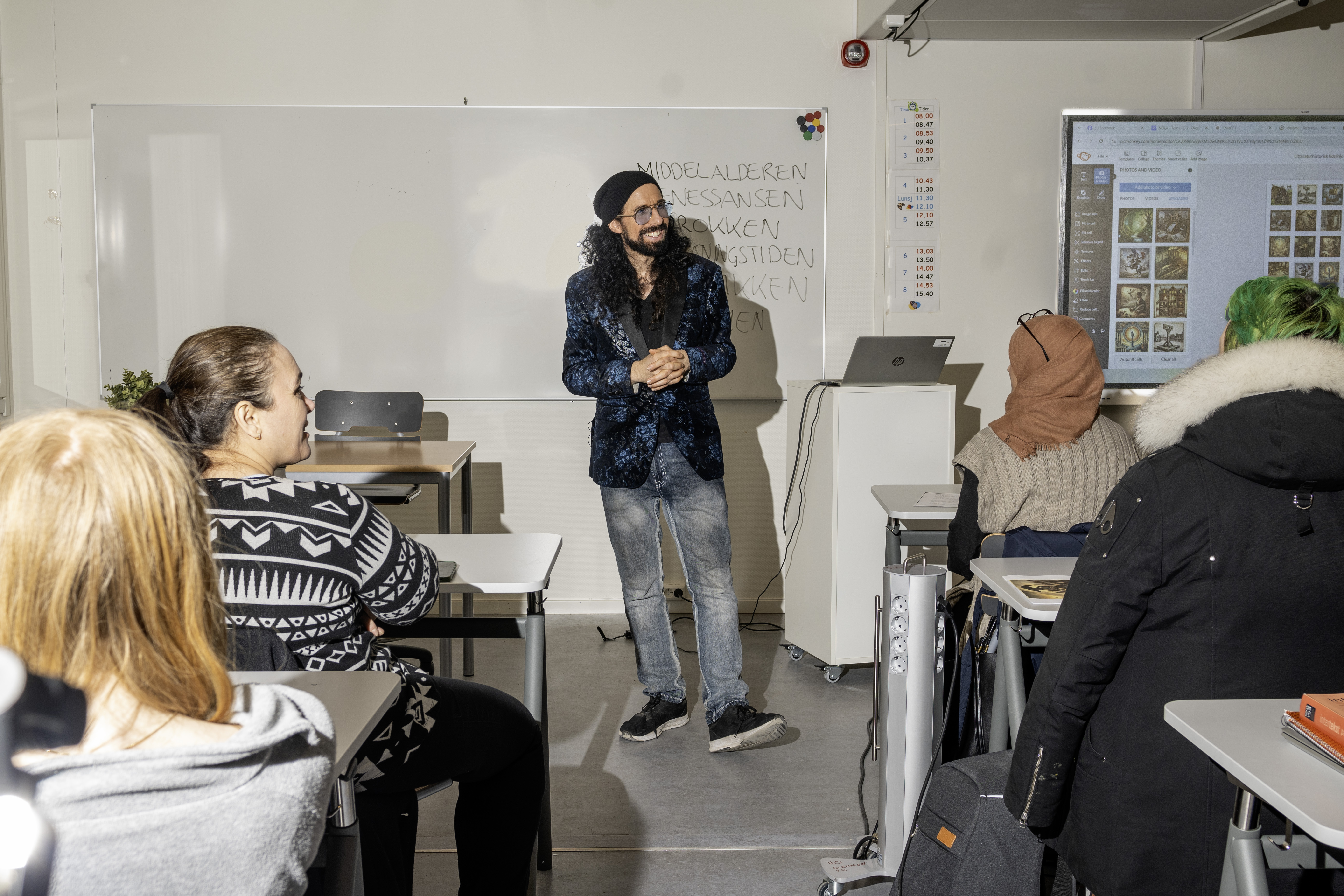 En mann står mellom elever som sitter i rader i et klasserom. Mannen har lange, mørke krøller og skjegg og ei svart, tettsittende lue på hodet. Han er kledd i mørkeblå, mønstrete dressjakke, olabukse og svarte sneakers. Foto.