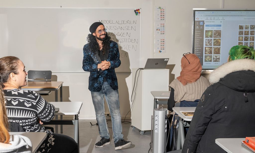 En mann står mellom elever som sitter i rader i et klasserom. Mannen har lange, mørke krøller og skjegg og ei svart, tettsittende lue på hodet. Han er kledd i mørkeblå, mønstrete dressjakke, olabukse og svarte sneakers. Foto.