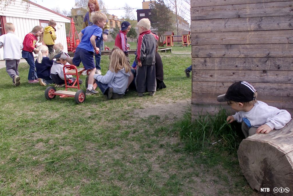 En liten gutt kryper sammen og kikker fram bak et hjørne. Han ser på at de andre barna leker i en barnehage. Foto.