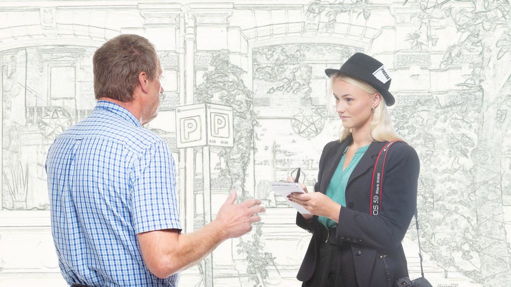 Kvinnelig journalist ikledd grå dress og hatt intervjuer en mann i blå- og hvitrutete skjorte. Foto.