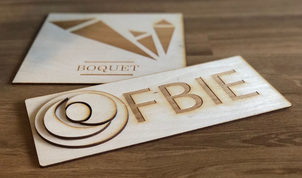 To treskilt med tekst og logo inngravert, det ene med teksten Boquet og det andre med teksten FBIE. Begge skiltene har stiliserte logoer bygget opp av dividerte former. Foto.