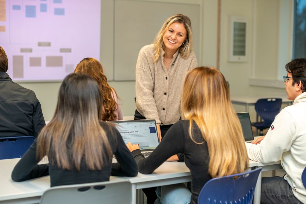 En kvinnelig lærer til smiler til elevene sine som jobber med skolearbeid på PC-ene sine i klasserommet. Foto.