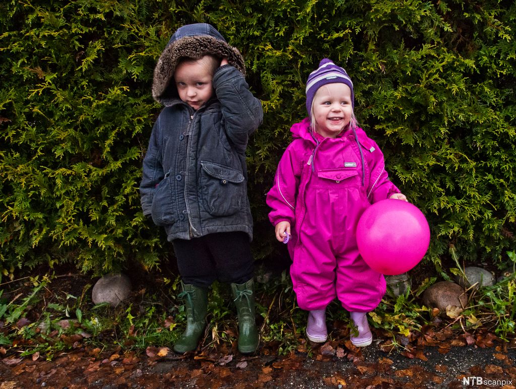 To små barn foran en hekk. Gutten har grønne støvler, svart bukse og mørk jakke. Han har hetta over hodet. Han kikker mot kameraet. Ved siden av står ei lita jente med rosa parkdress, lilla lue og lilla støvler. Hun holder en stor rosa ballong, smiler og ser mot noe bortenfor seg. Hun holder en smokk i hånda. Foto.