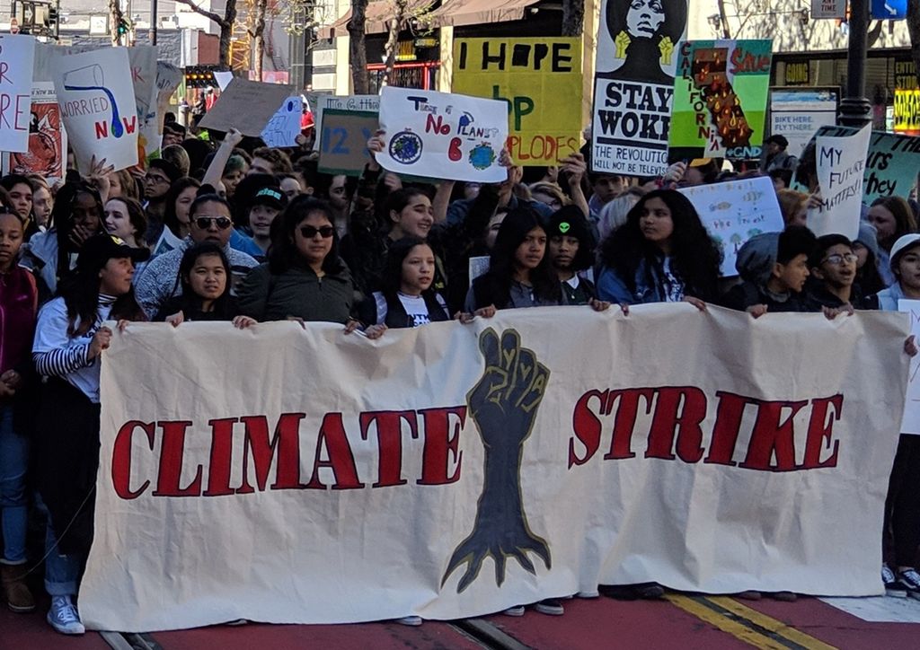 Large banner with a clenched fist with roots and the words Climate Strike, many young people walking in protest behind the banner. Photo.