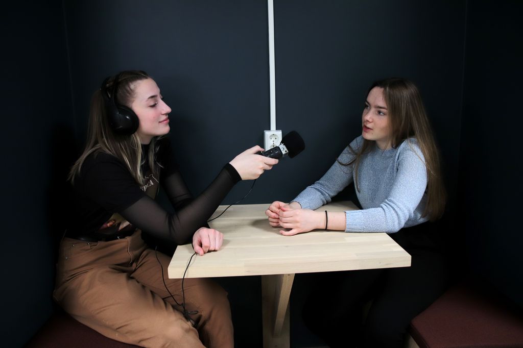 To jenter sitter ved et bord. Jenta til venstre har øreklokker og lydopptaker og lytter til jente til høyre som snakker. Foto.