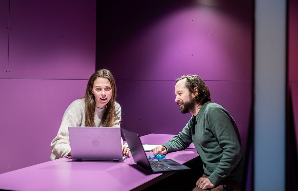 To personer, en dame og en mann, sitter i et lilla rom med hver sin laptop. Foto.