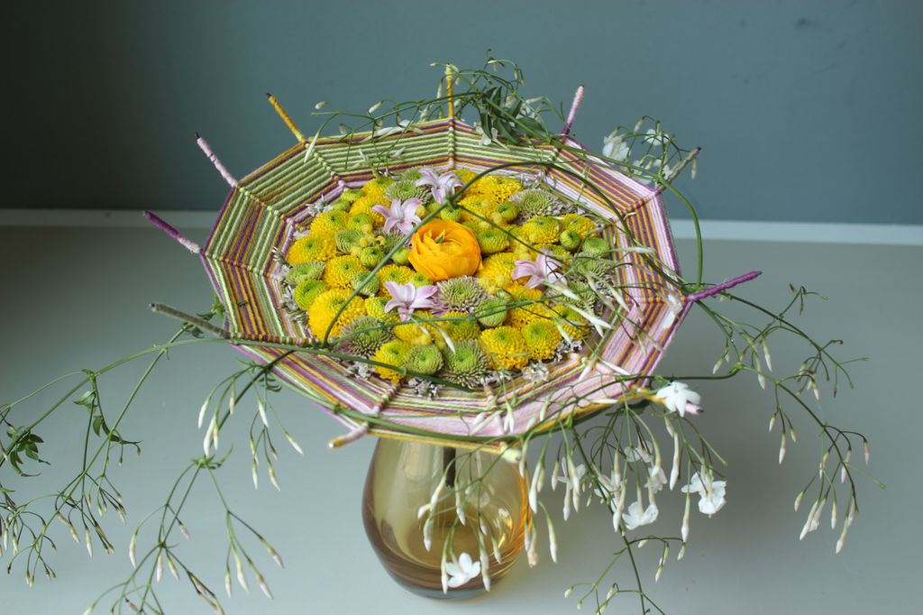 Stativ laget av ståltråd som et spindelvev, vevd med garn i ulike farger og blomster. Foto.