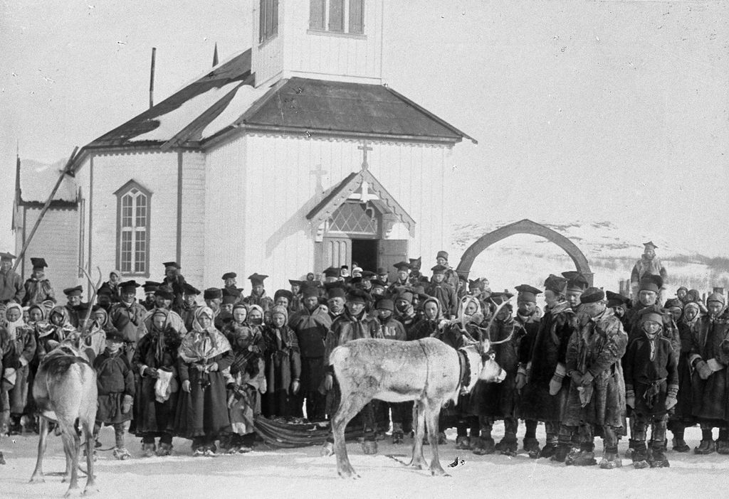 En stor gruppe mennesker, de fleste i samiske drakter, og et par
 reinsdyr foran en kirke. Bildet er tatt vinterstid. Svart-hvitt foto. 