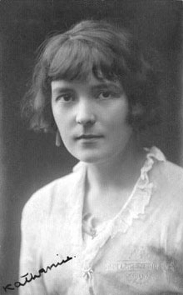 Photo: Black and white portrait of Katherine Mansfield. We see a woman with dark hair, and dark eyes. She is wearing a white blouse with a frilly collar. She looks serious. 