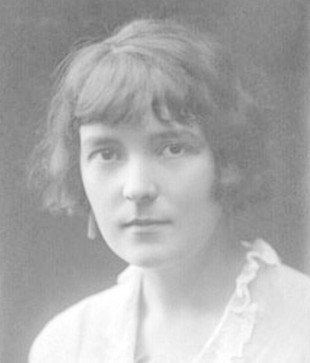 Photo: Black and white portrait of Katherine Mansfield. We see a woman with dark hair, and dark eyes. She is wearing a white blouse with a frilly collar. She looks serious.