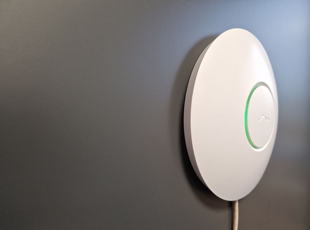 Hvit tallerkenformet enhet som henger på en vegg. En nettverksledning går ned fra enheten. Foto.