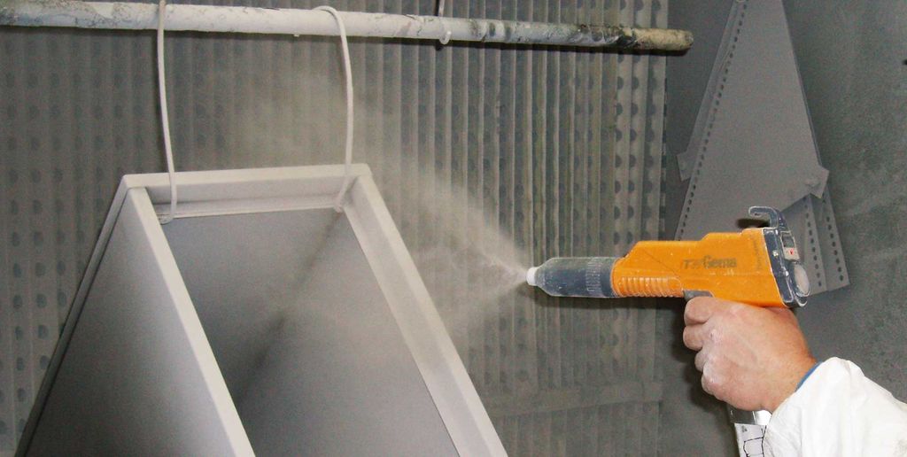 Hånd som holder en pulverlakkeringspistol som sprayer pulver på en metallgjenstand i en lakkboks. Foto.