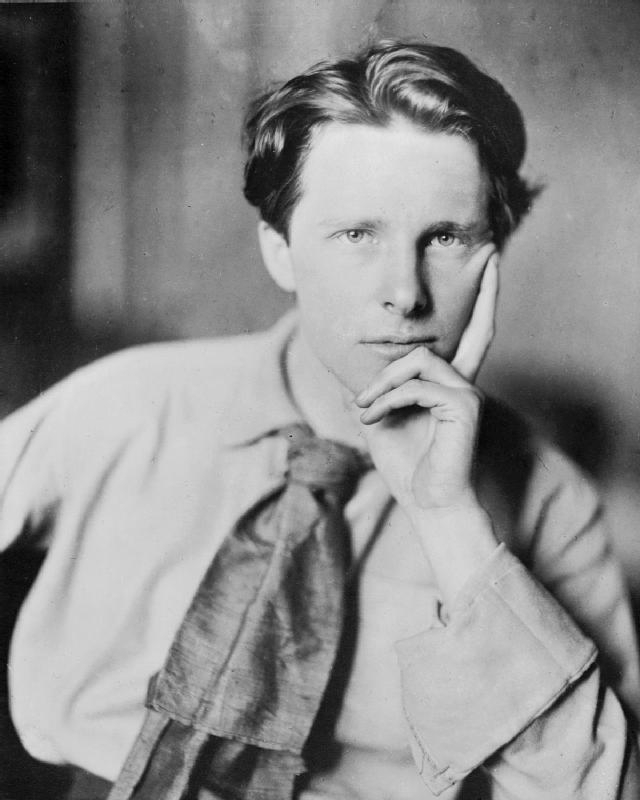 Black and white portrait of Rupert Brooke. He is a young man looking directly at the camera. He is wearing a white shirt, and a scarf tied as a tie. 
