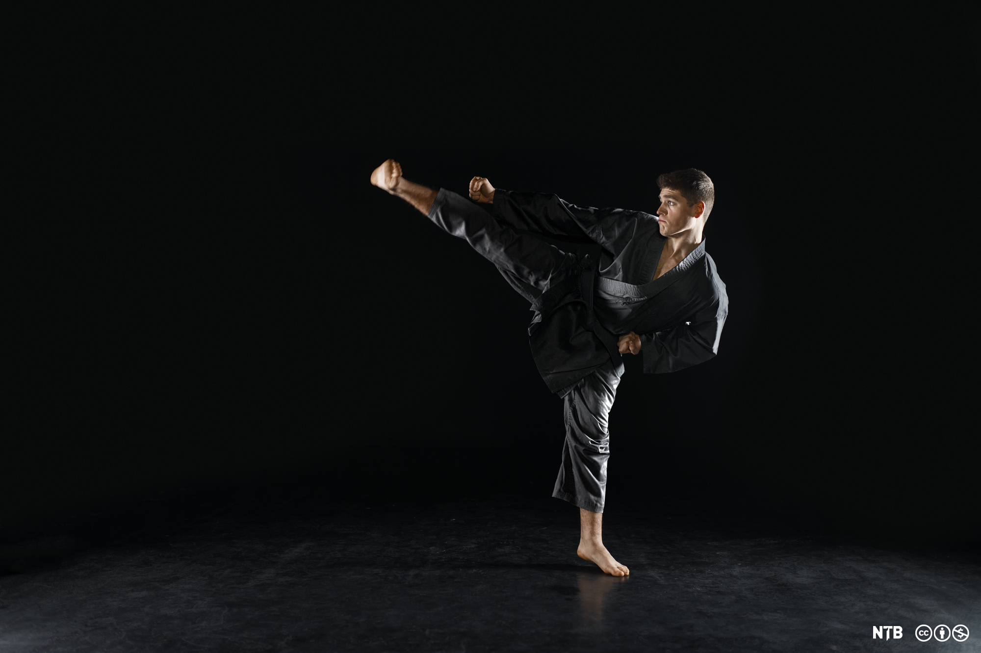 Mann med svart karatedrakt står på eitt bein og held det andre beinet svært høgt opp. Foto. 