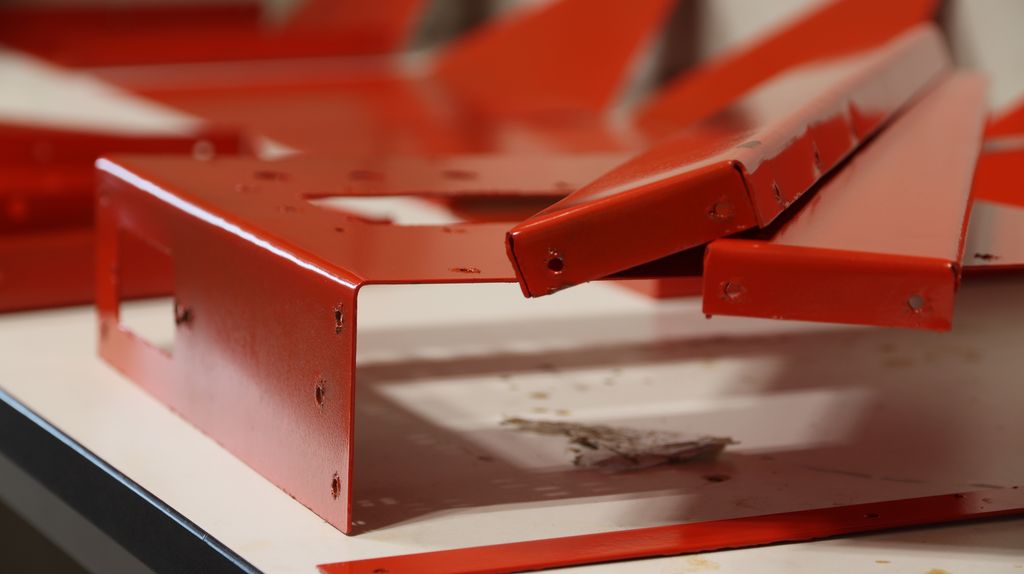 Rødlakkerte, bøyde metalldeler liggende på et bord. Langsmed kantene på delene er det stansa ut små hull. Foto.