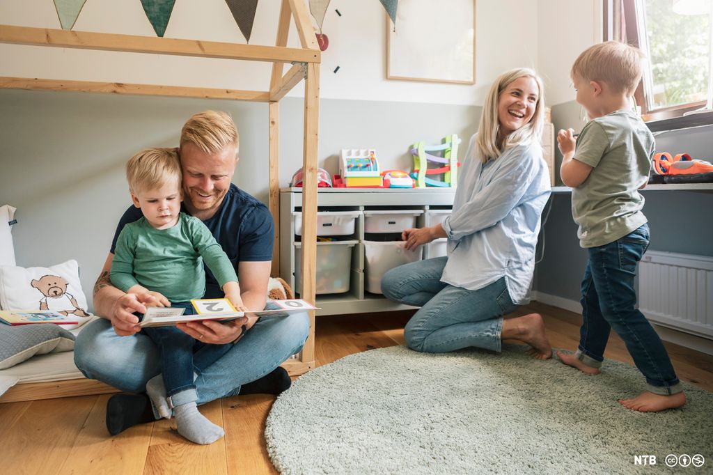 To voksne og to barn på et barnerom. En gutt sitter på fanget til en mann, de leser i ei bok. Ei kvinne hjelper en gutt med å rydde. Foto.