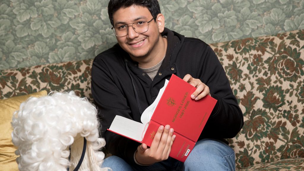 En ung mann sitter på en sofa og holder boka Norges lover i hendene. Foran ham er det en parykk med hvitt, krøllete hår på et stativ. Foto.