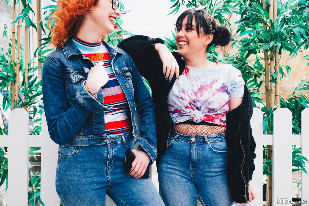 To unge smilende kvinner foran et gjerde og planter. Foto. 