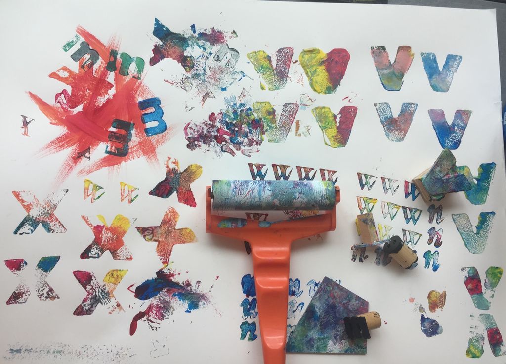 Et hvitt ark som er fylt med bokstavtrykk i ulike farger. Oppå arket ligger det en sjablong og ei rulle. Foto.