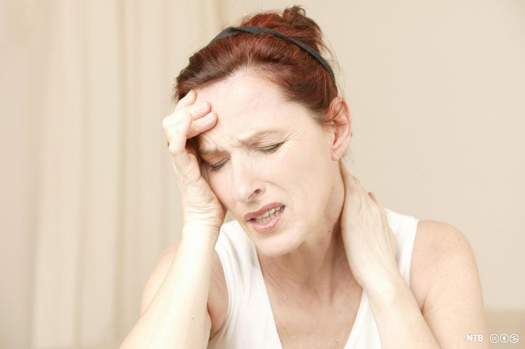 Middelaldrende kvinne som holder en hånd i nakken og den andre ved tinningen. Hun har et plaget uttrykk i ansiktet. Foto.