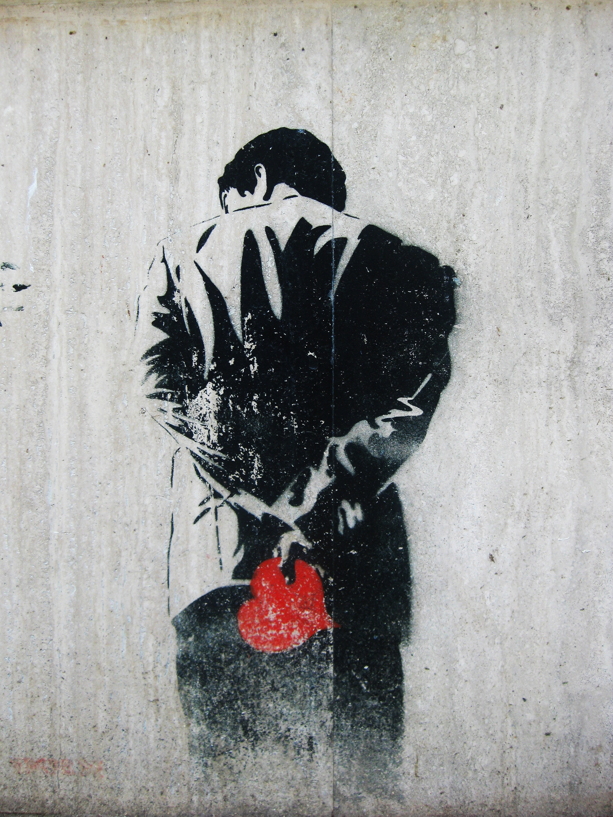 Graffiti på en murvegg. En person står med ryggen til. Han har hendene på ryggen og i den ene hånda holder han et rødt hjerte. Foto.