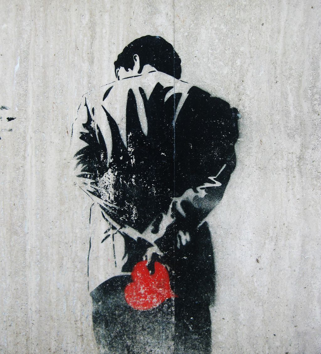 Graffiti på en murvegg. En person står med ryggen til. Han har hendene på ryggen og i den ene hånda holder han et rødt hjerte. Foto.