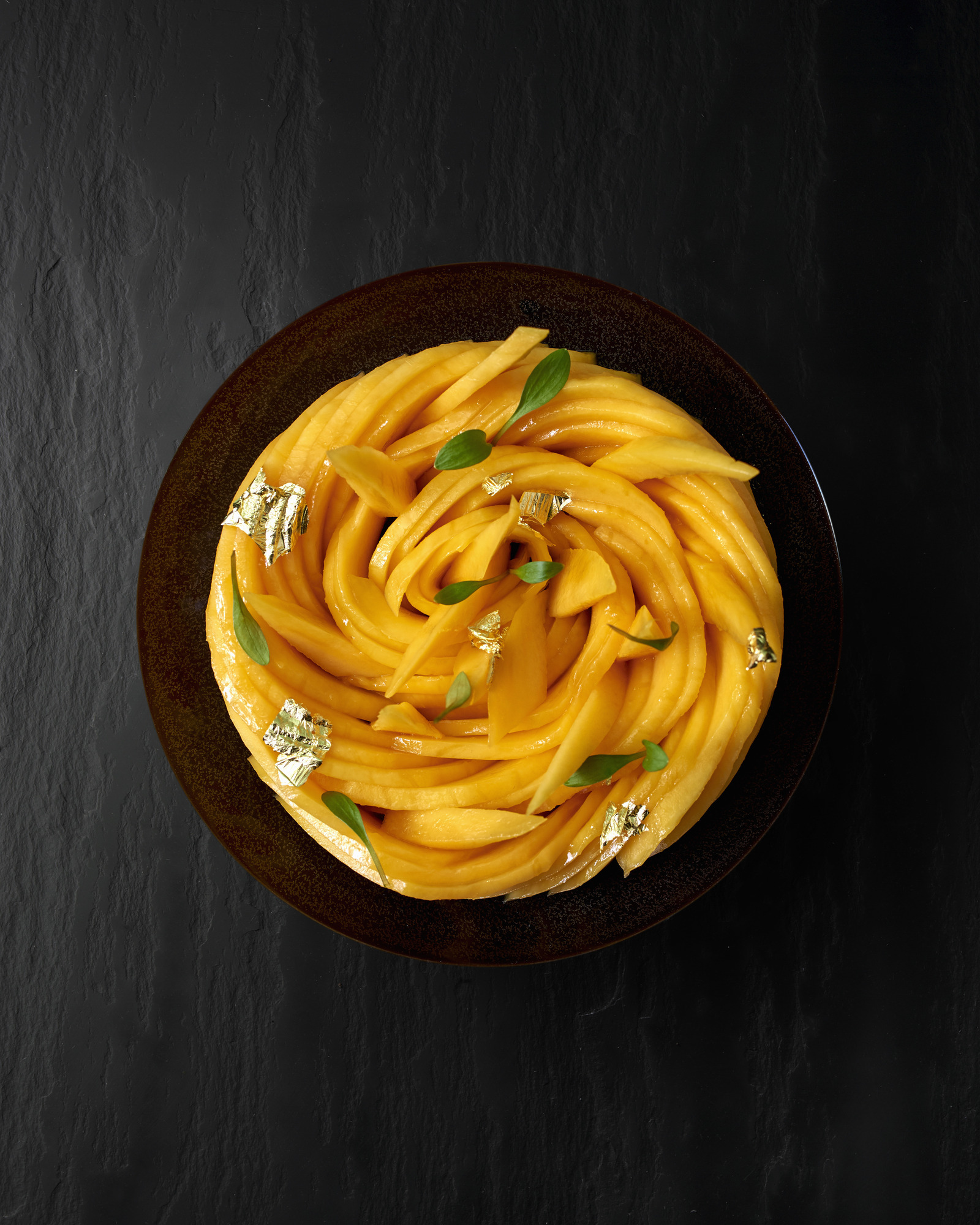 Skål med tynne skiver av mango lagt slik at de danner en sirkulær form. Grønne blader og blader av gull er brukt som garnityr. Foto.