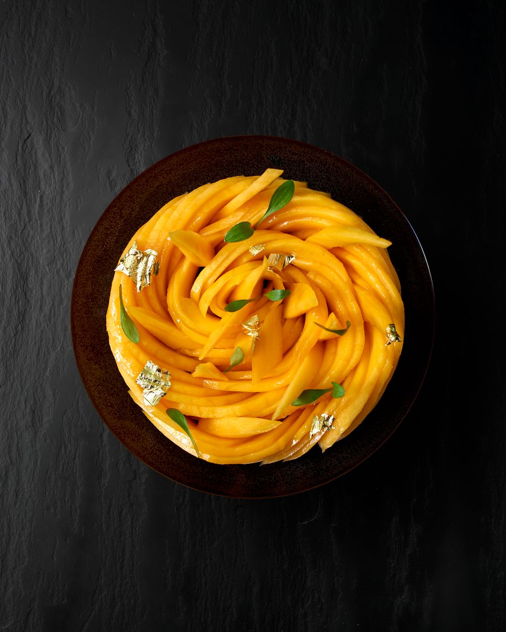 Skål med tynne skiver av mango lagde slik at dei dannar ei sirkulær form. Grøne blad og blad av gull er brukte som garnityr. Foto.