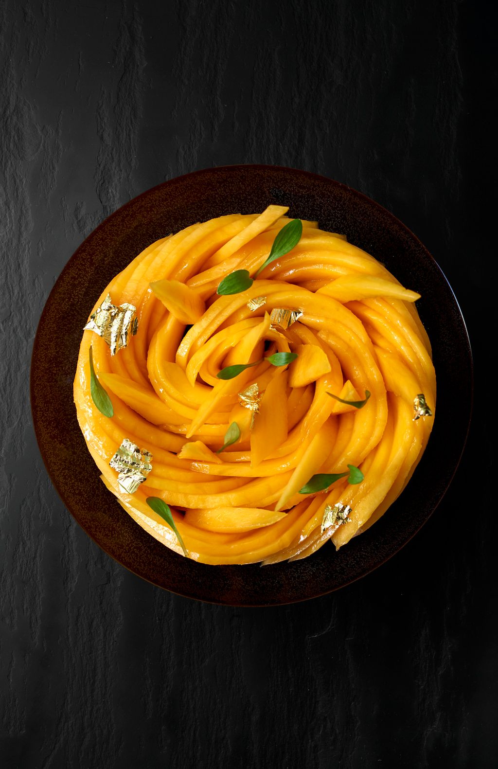 Skål med tynne skiver av mango lagt slik at de danner en sirkulær form. Grønne blader og blader av gull er brukt som garnityr. Foto.