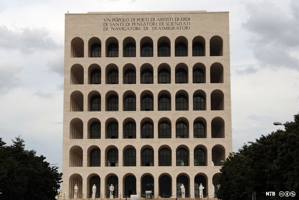 Bygningene er høy, den har seks etasjer med ni like rundbuede arkader foran vinduene som ligger dypere inn. Øverst er det et bredt felt med skrift. På gateplan står det hvite statuer. Foto. 