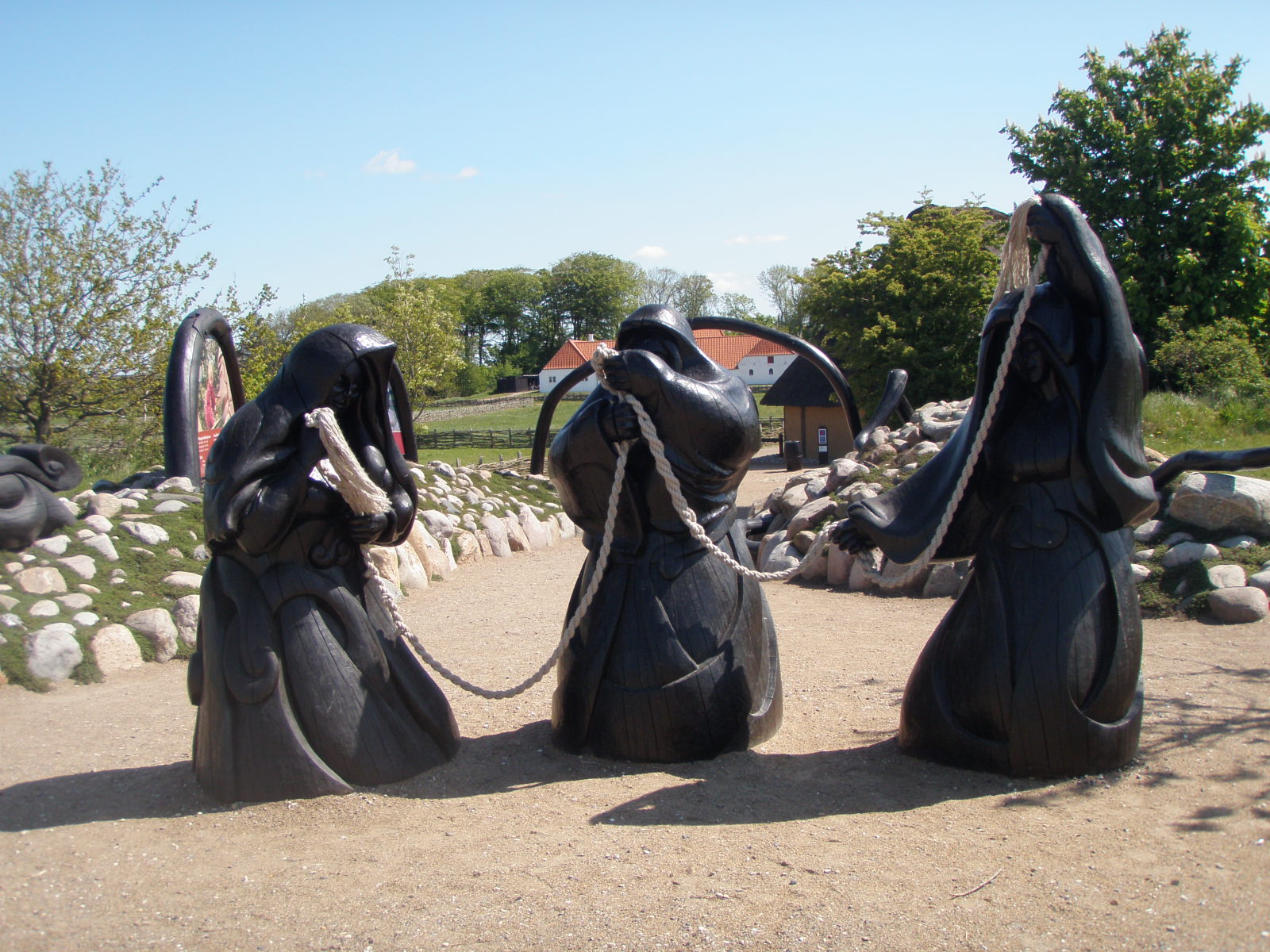 Statuer av tre sortkledde kvinner som holder et tau står på et fast underlag. I bakgrunnen ser vi mark, trær og hus. Foto.
