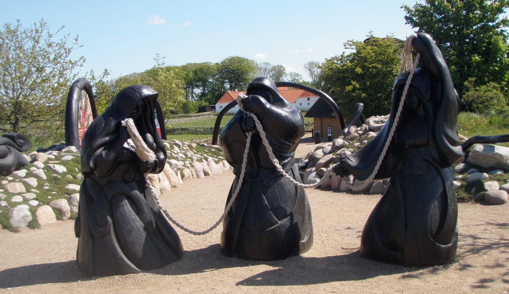 Statuer av tre sortkledde kvinner som holder et tau står på et fast underlag. I bakgrunnen ser vi mark, trær og hus. Foto.