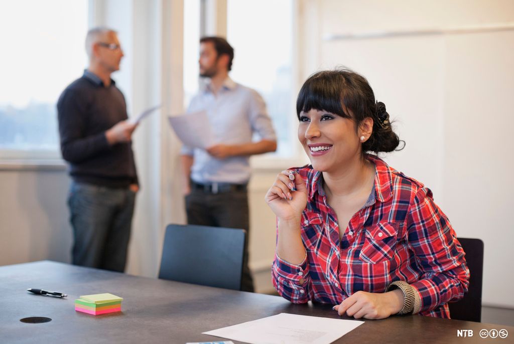 Smilende kvinne sitter ved et møtebord, to menn som holder papirer, står i bakgrunnen. Foto.