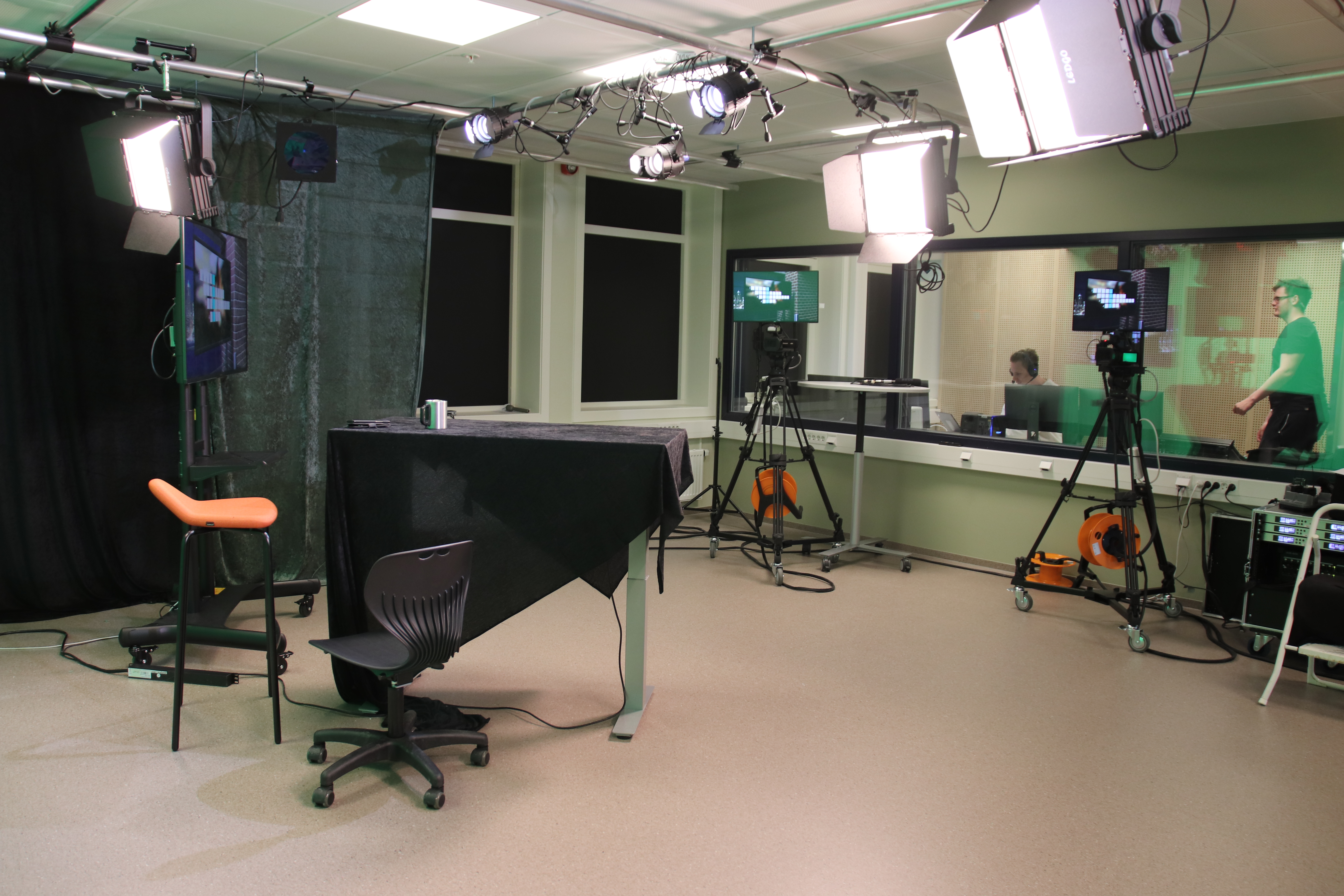 Tv-studio med dukkledd bord og stoler til programleder, flere kameraer og skjermer og flere lyskastere i taket. To personer i kontrollrommet i bakgrunnen. Foto. 