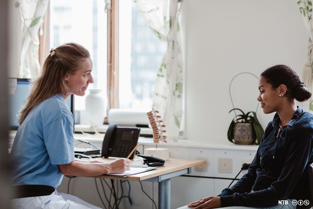 Ei ung jente sitter og snakker med en helsesykepleier på et kontor. Foto