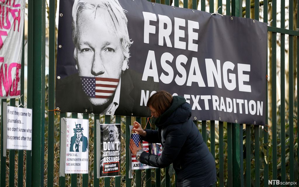 Ei kvinne festar ein plakat på eit gjerde der det heng ein stor "Free Assange"-poster. Foto.